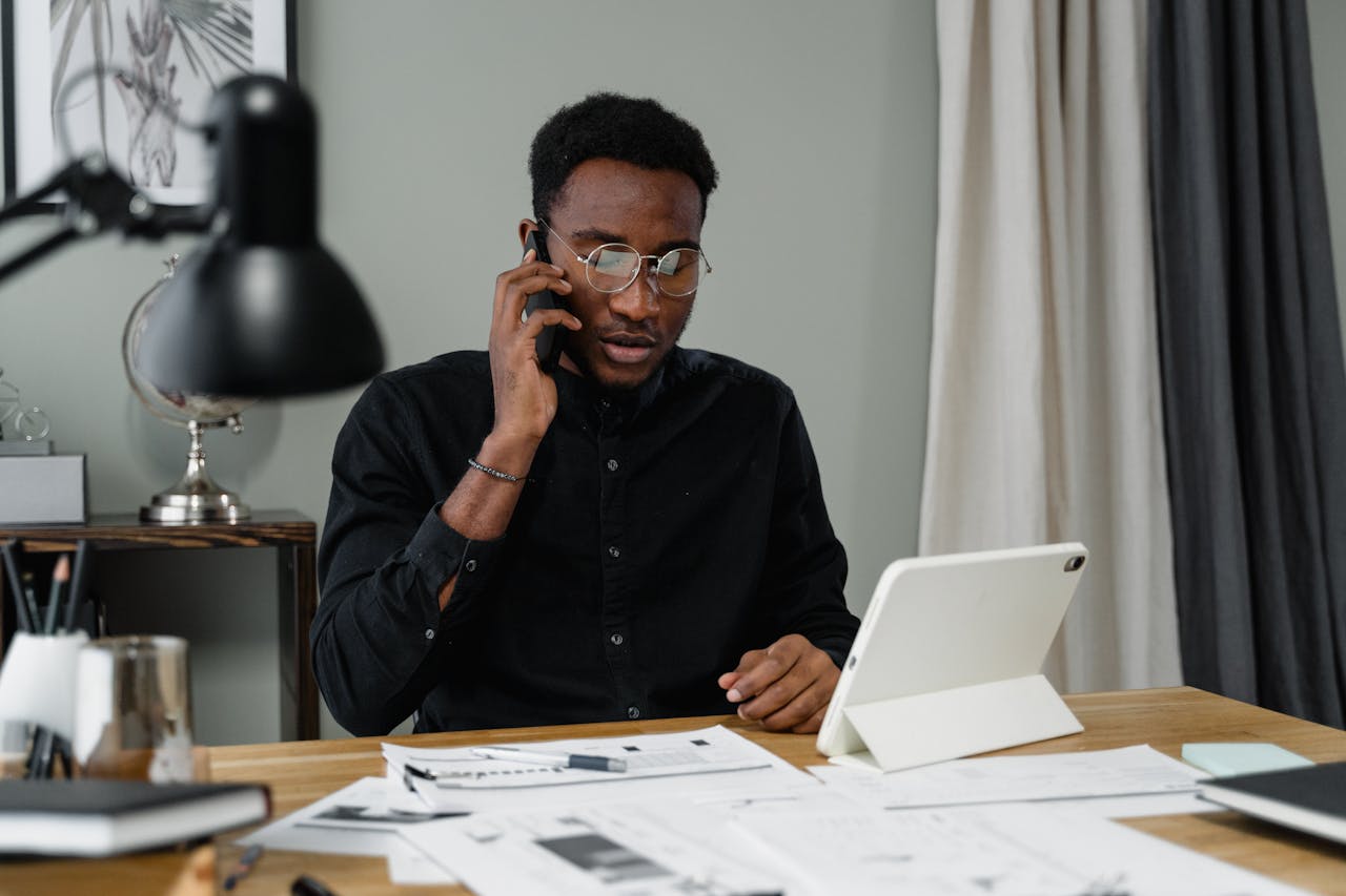 Focused man with eyeglasses making a work-related phone call at his desk with documents and a tablet.