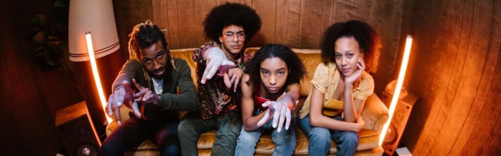 Four young friends pose energetically while sitting on a vintage couch indoors.