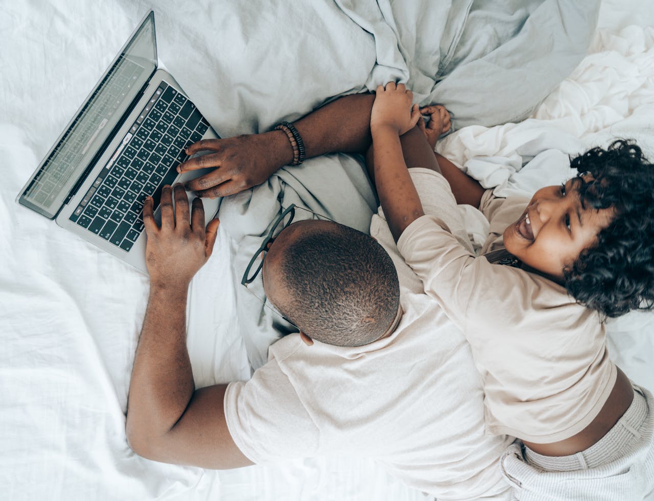 Top view of cheerful African American kid lying near father while man typing on keyboard of laptop during remote work in cozy bedroom