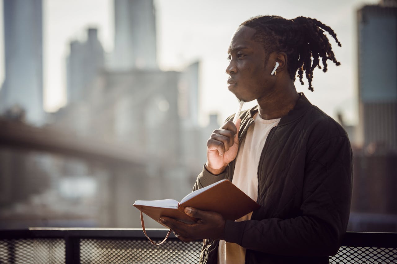 Man with earphones and notebook pondering outdoors, cityscape in background.